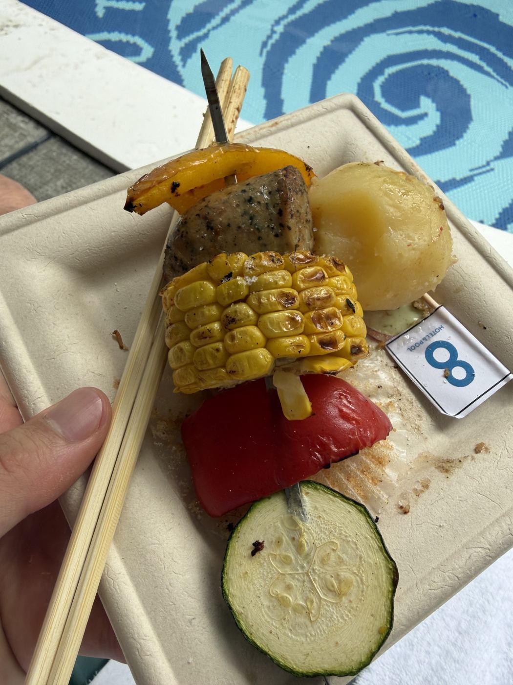 プールサイドで食べるBBQ串。焼きとうもろこしと野菜、8HOTELの旗が添えられている