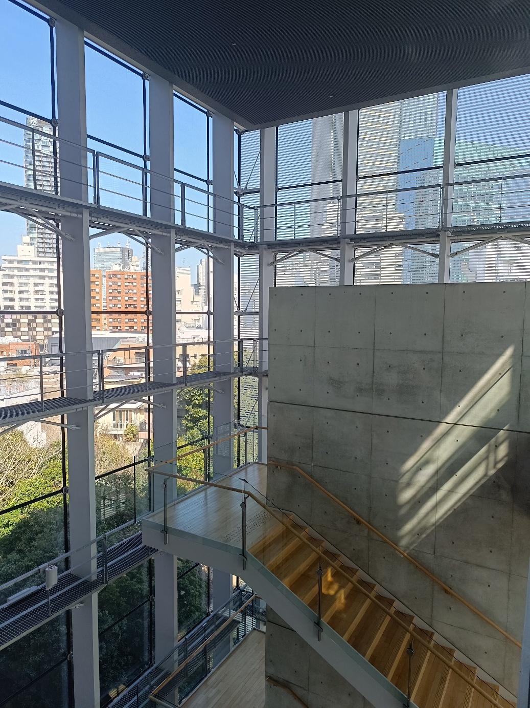 Interno del National Art Center di Tokyo: la vetrata a tutta altezza inquadra i palazzi di Roppongi, accanto a un muro di cemento a vista e a una scala in legno
