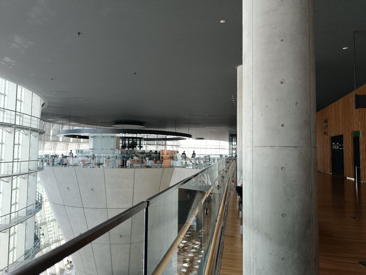 Ballatoio interno del National Art Center di Tokyo con pavimento in legno e un pilastro di cemento, affacciato sull'atrio e sul cono del ristorante